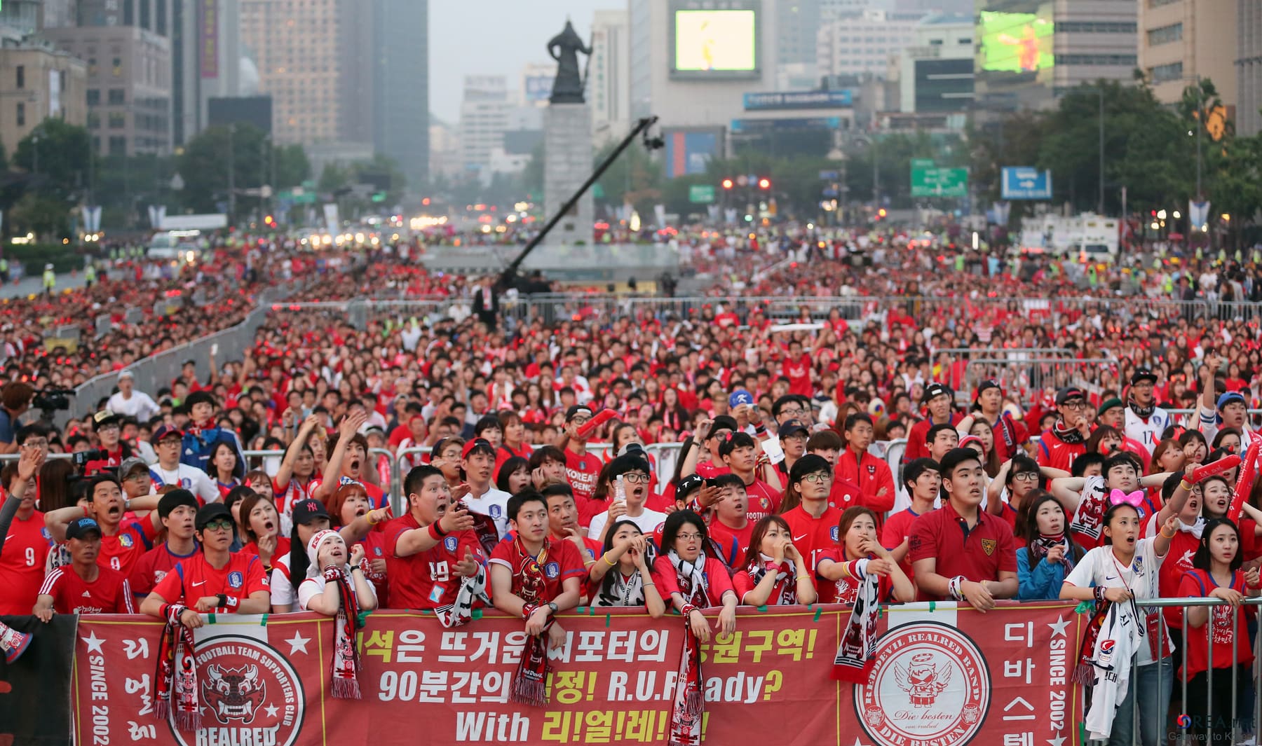 A packed crowd of fans in red shirts cheering in central Seoul beneath Hangul banners