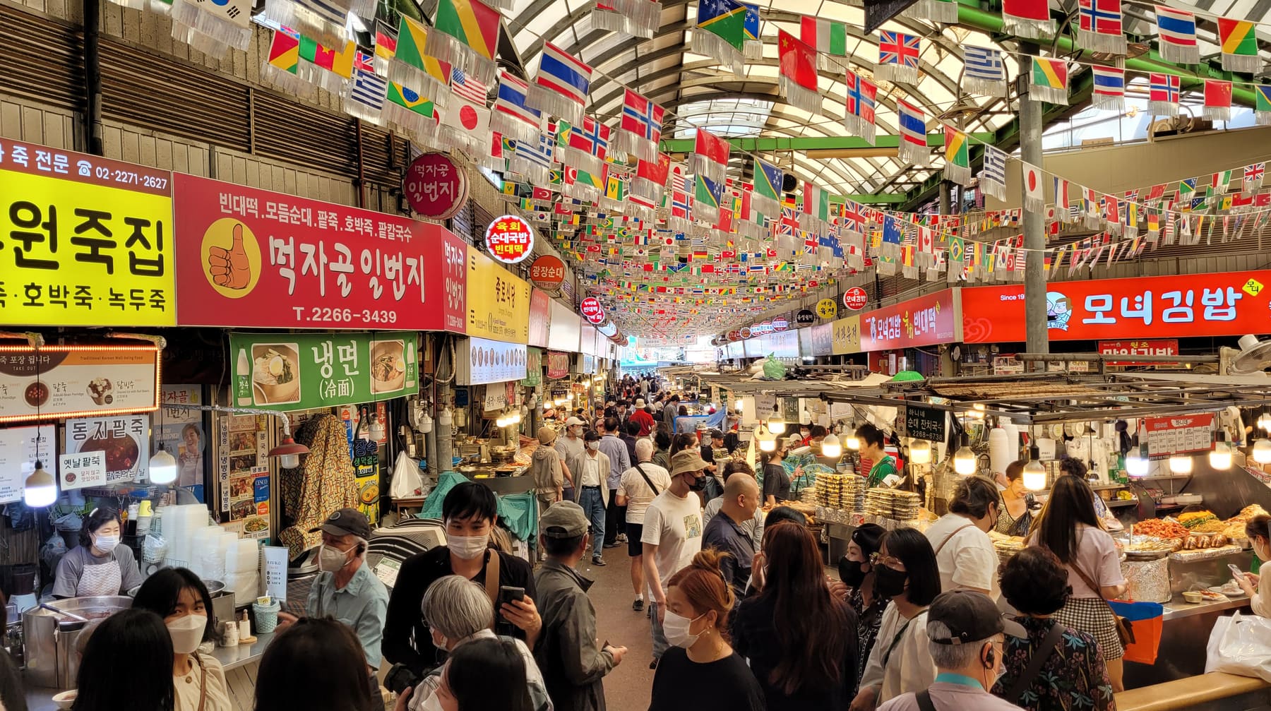 A crowded covered aisle at Gwangjang Market in Seoul under hanging flags, with Hangul shop signs