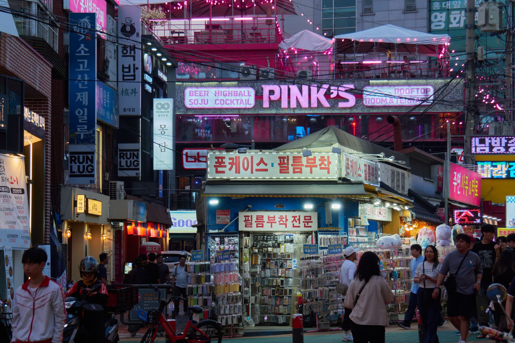 A busy evening street in the Hongdae area of Seoul with neon Hangul signs and passers-by