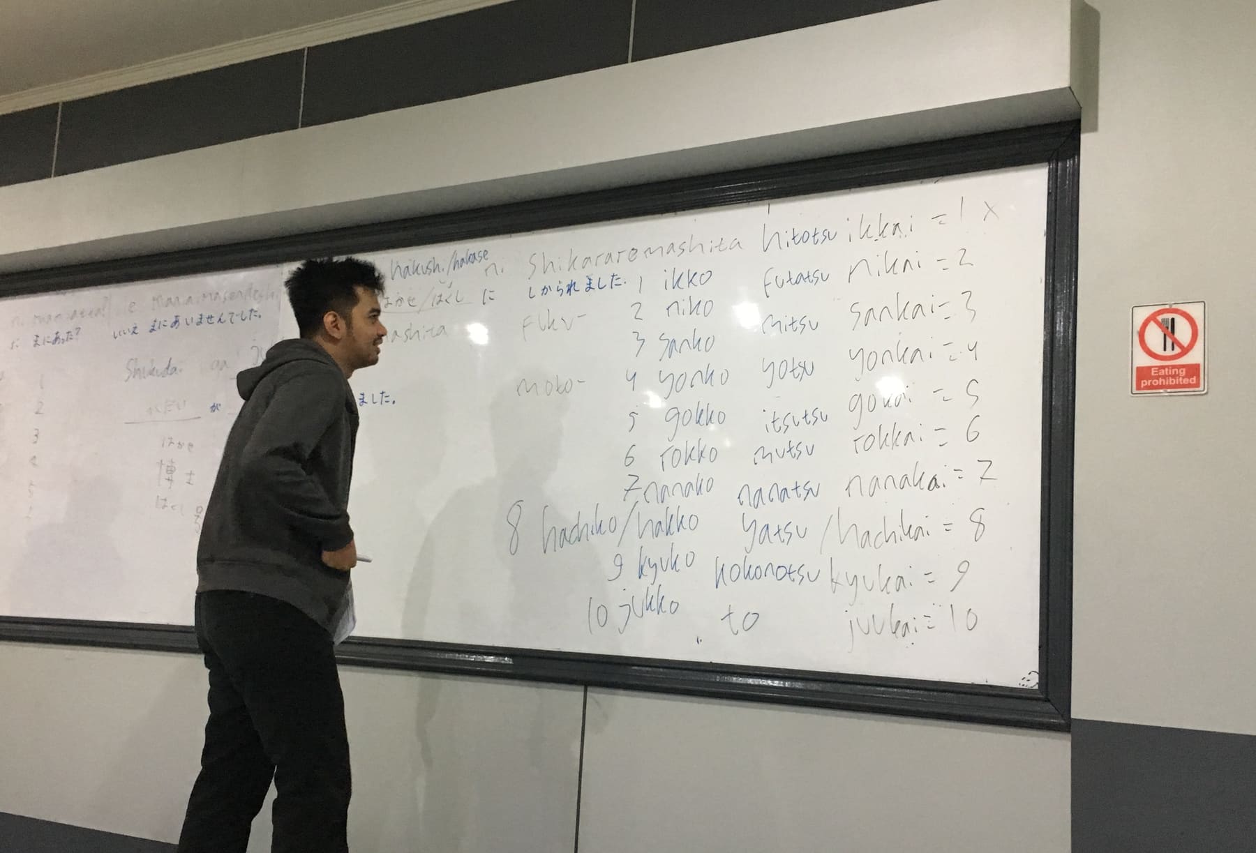 A man writing numbers in romaji on a large whiteboard in a Japanese lesson