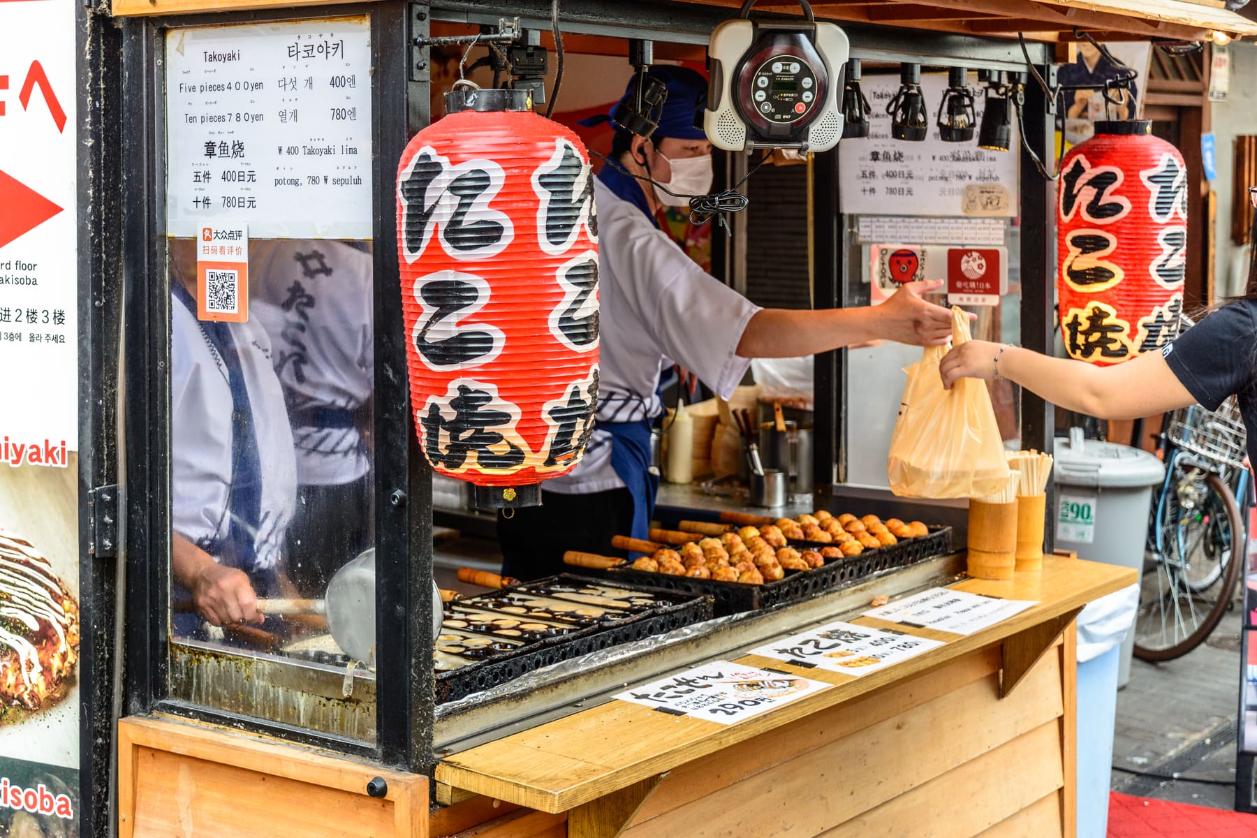 A vendor at a takoyaki stall passing a paper bag across the counter to a customer